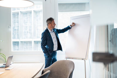 Businessman with hand in pocket writing on flipchart at office