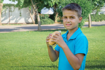 Happy boy in a blue t-shirt eats a hamburger outdoors in the park.