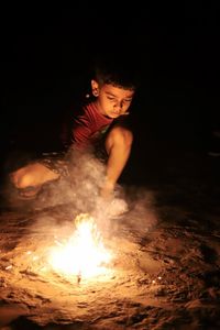 Cute boy crouching by fireworks on field at night