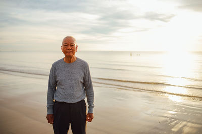 Portrait of senior man standing at beach against sky during sunset