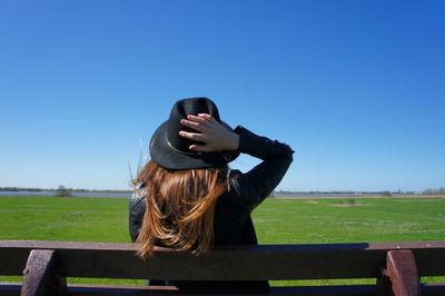 Rear view of woman wearing hat sitting on bench at field against clear blue sky during sunny day