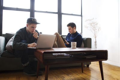 Man using laptop while friend reading book at home