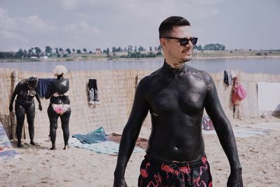 Man with black mud on body standing at beach against sky