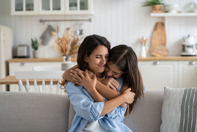 Loving bored girl hugs woman sits on couch at home clinging to from behind feeling affection