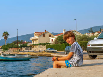 A boy in a striped t-shirt and swimming trunks is sitting near the seashore and looks at the camera