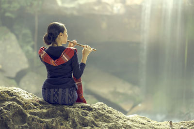 Rear view of woman playing flute while sitting on rock against waterfall