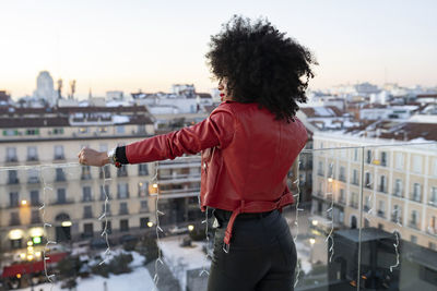 Back view of african american female with curly hair in stylish outfit standing on balcony and looking at city buildings