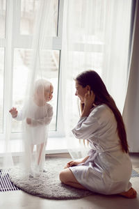 Mother in a white robe sits with a child a blonde daughter at a large window of the house person