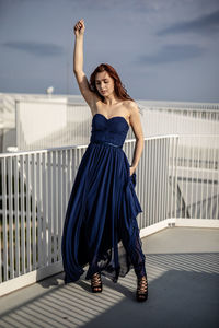 Woman in dress with hand raised standing in balcony against sky