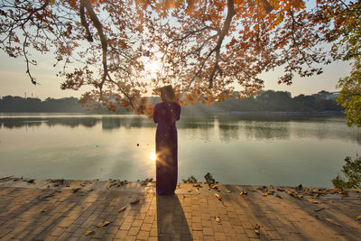 Rear view of woman standing by lake against sky during sunset