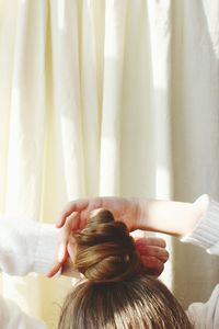 Cropped image of woman tying hair bun against curtain at home