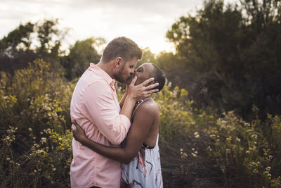 Side view of romantic couple kissing while standing amidst plants against sky in park during sunset