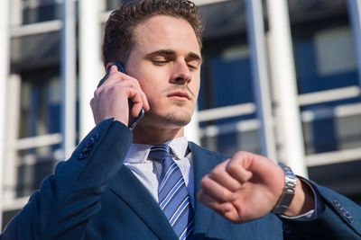 Low angle view of businessman talking on phone while checking time in city