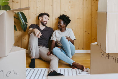 Happy multiracial couple talking while sitting together near wooden wall at new home