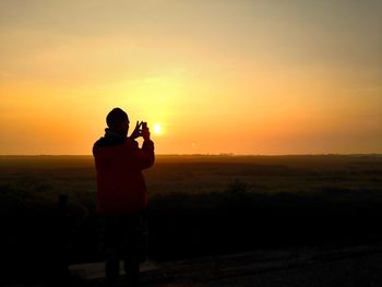 Silhouette man photographing on field using smart phone against sky during sunset