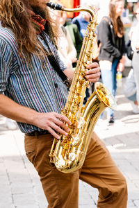 Vertical photo of the hands of a young man with long hair playing saxophone in a crowded the street