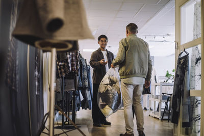 Rear view of male fashion designer giving plastic bag of recycled clothes to colleague at workshop