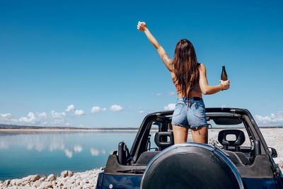 Back view of excited woman in summer outfit raising hand of beer while standing on roof of safari car on shore of river