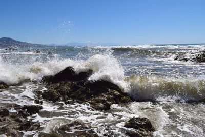 Sea waves splashing on rocks at shore against clear blue sky