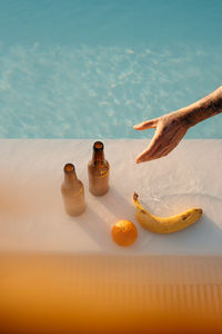 From above of crop unrecognizable tattooed tourist reaching out hand towards bottles of beer placed near swimming pool with fresh banana and orange during holidays in mallorca person