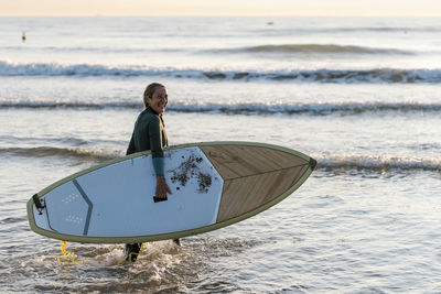 Woman laughing while holding paddleboard standing in sea during dawn