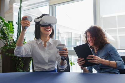 Two women sitting at the table in the office and working with virtual reality viewer and tablet