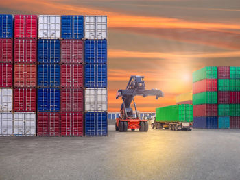 Vehicles by cargo containers at shipyard against sky during sunset