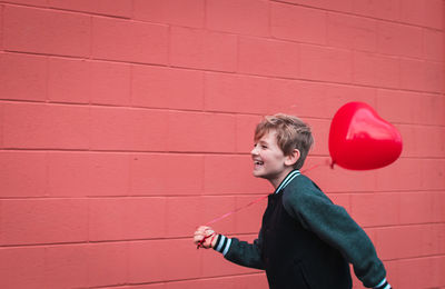 Smiling boy holding balloon while walking by red wall