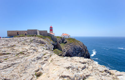 Lighthouse on rocks by sea against clear blue sky