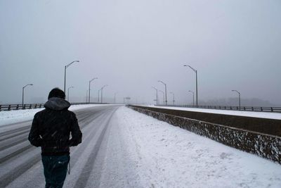 Rear view of man walking on road against sky during snowfall