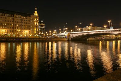 Illuminated bridge over river by buildings against sky at night