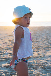 Little blonde girl in a panama hat stands with her back on the beach and looks at the ocean