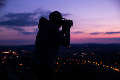 Silhouette man photographing illuminated cityscape against sky during sunset