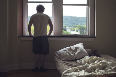 Rear view of man looking through window from bedroom