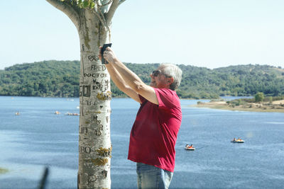 Senior man photographing while standing by sea against clear sky