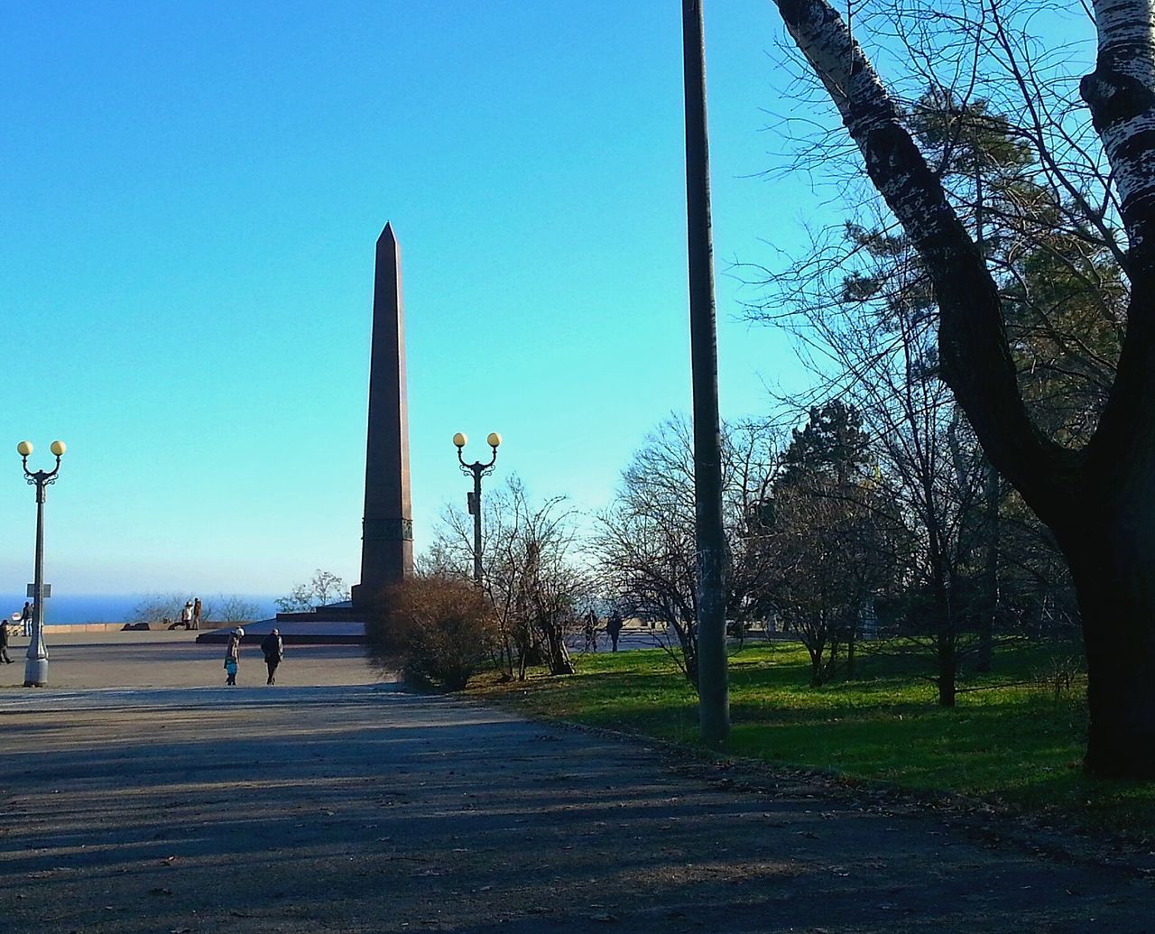 clear sky, tree, blue, street light, sunlight, built structure, grass, footpath, shadow, the way forward, road, architecture, incidental people, sky, park - man made space, day, building exterior, copy space, outdoors, street