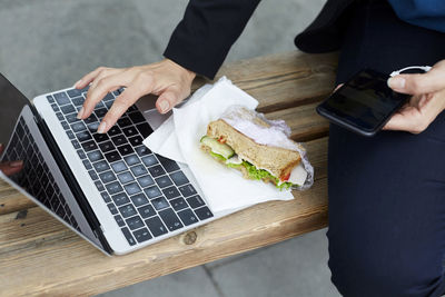 Midsection of businesswoman using laptop while sitting with smart phone and sandwich at bus stop in city