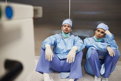Full length of tired surgeons sitting on floor in hospital