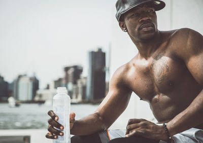 Midsection of shirtless man drinking glass against sky in city