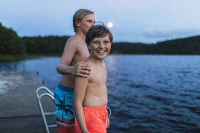 Portrait of smiling shirtless boy standing with brother by sea at dusk