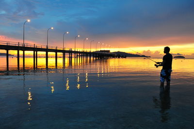 Silhouette man fishing in water against orange sky