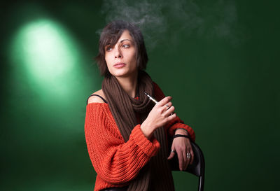 Young woman using electric cigarette while sitting against green background