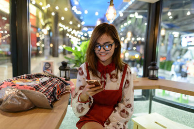 Smiling young woman using phone while sitting in restaurant