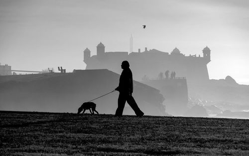 Silhouette man with dog walking against castle during foggy weather