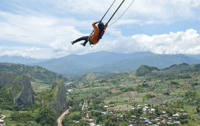 Woman swinging over landscape against cloudy sky