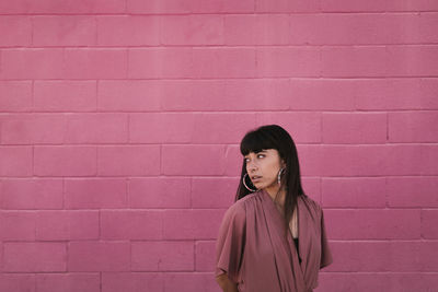 Side view of stylish young ethnic female with long dark hair in trendy dress standing against pink wall on street and looking away thoughtfully