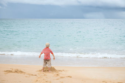 Rear of little boy with hat play sand on beach with motion sea wave, phuket