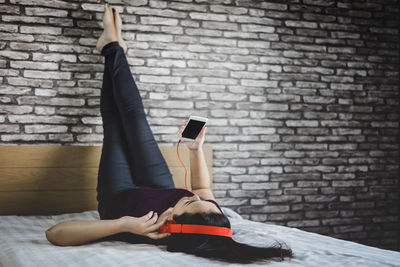 Woman using mobile phone while lying on bed against brick wall at home