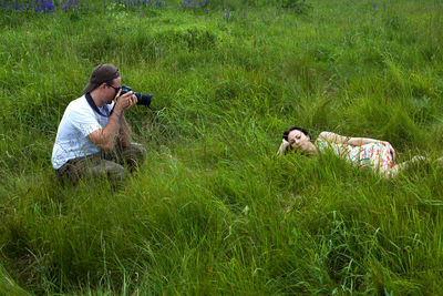 Side view of man photographing woman lying on grassy field