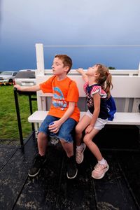 Siblings sitting on chair at shore against sky